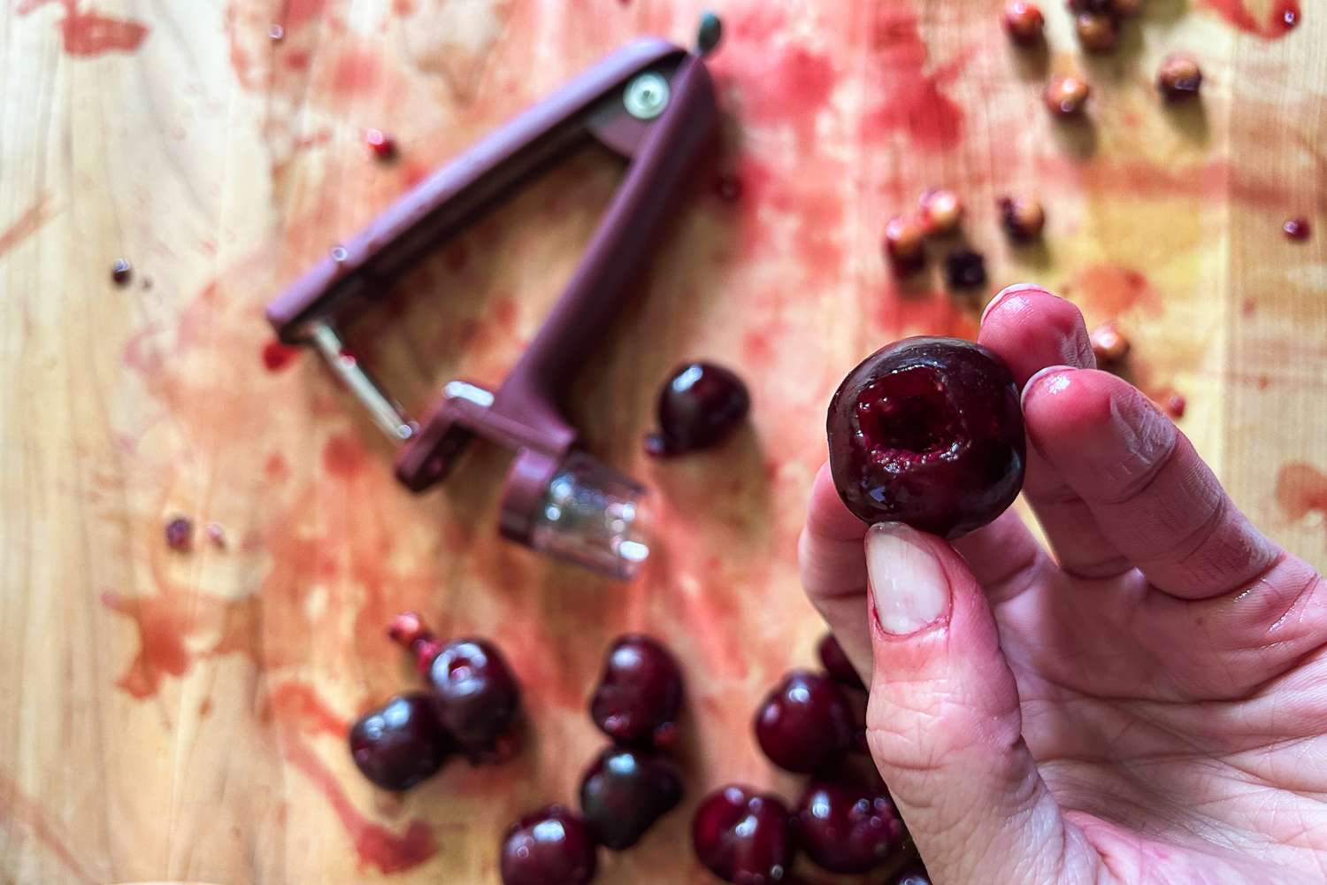 A closeup of a pitted cherry with a juice-stained cutting board and cherry pitter in the background