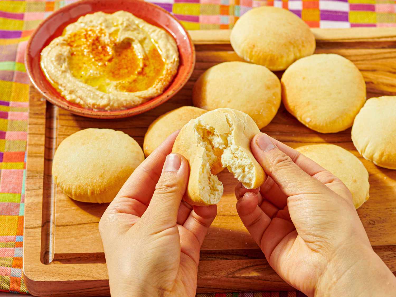Hands holding a torn piece of pita bread wooden board with pita and a bowl of hummus in the background