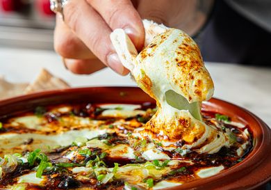 A hand dipping a piece of bread into a baked cheese and herb dish served in a terracotta bowl