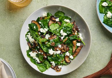 Warm kale and caramelized mushroom salad in a white ceramic bowl on a green surface. There are a pair of wooden spoons at the bottom right corner, a glass of golden liquid in the top left corner, a stack of plates covered with a napkin in the bottom left corner, and another plate of salad in the top right corner.