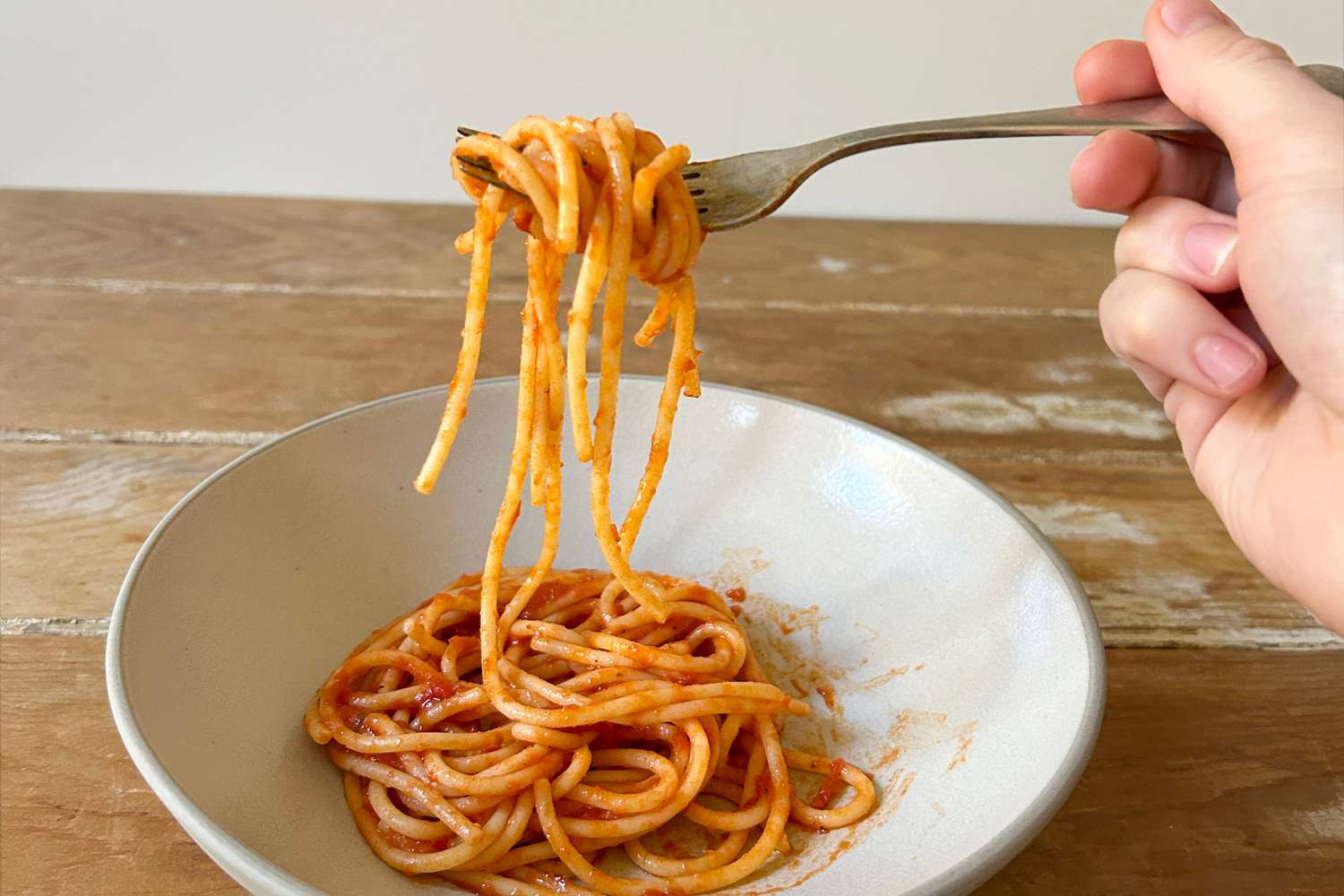 A hand holding a forkful of spaghetti with red sauce above a white pasta bowl.