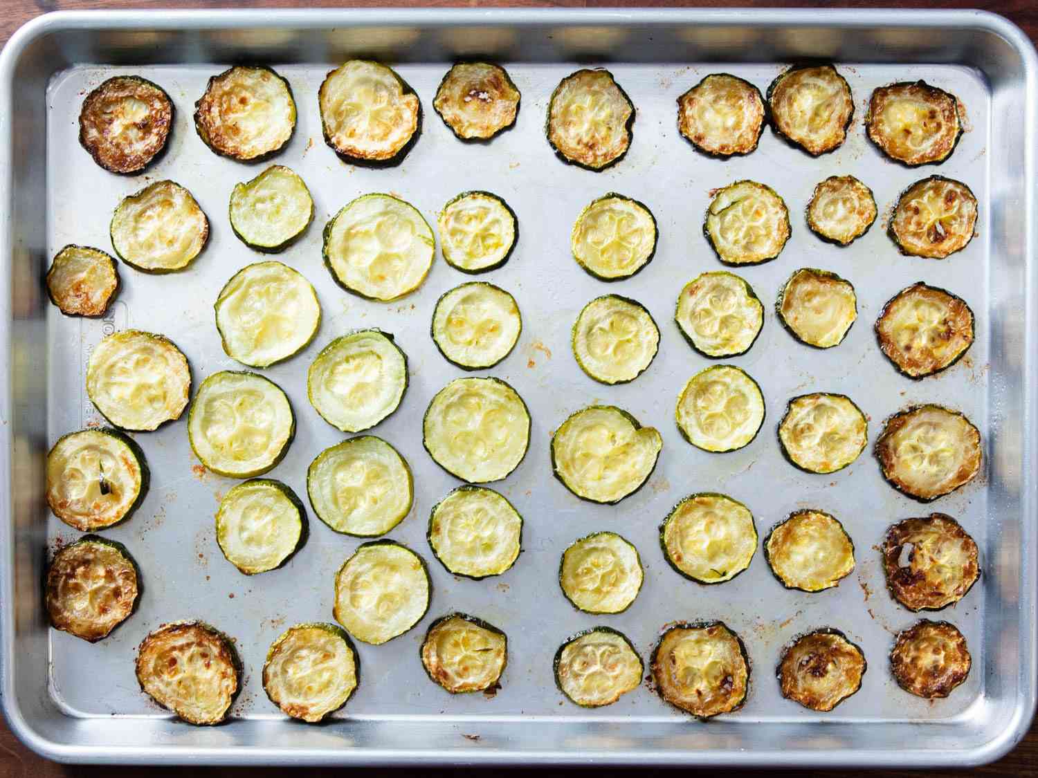 Zucchini rounds spread evenly over a baking sheet: the ones on the perimeter are much more darkly browned than the ones in the center, showing how heat is not evenly distributed on a baking sheet.