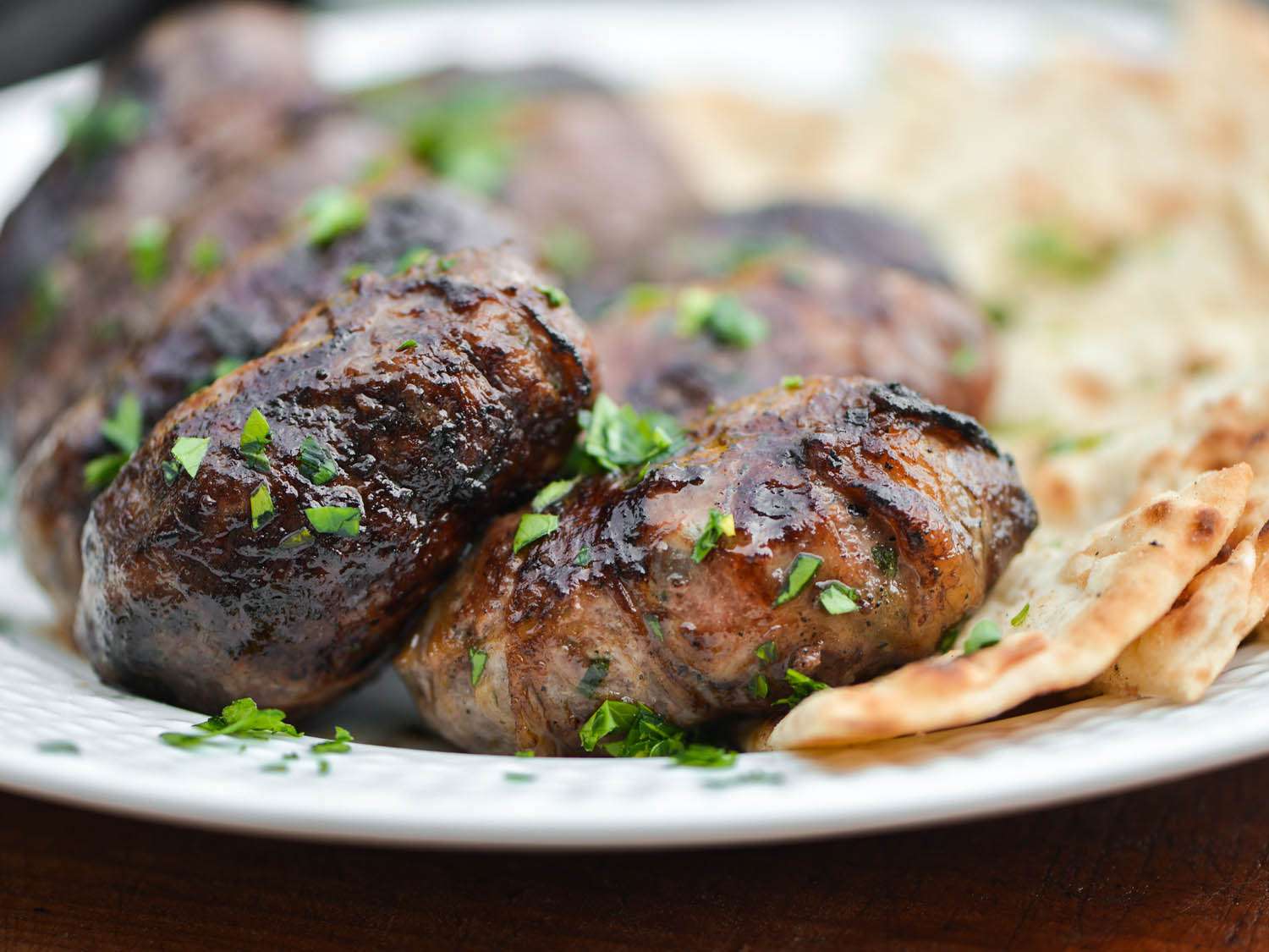 Close-up of grilled sheftalia sausages on a plate, showered with parsley and flanked by flatbreads.