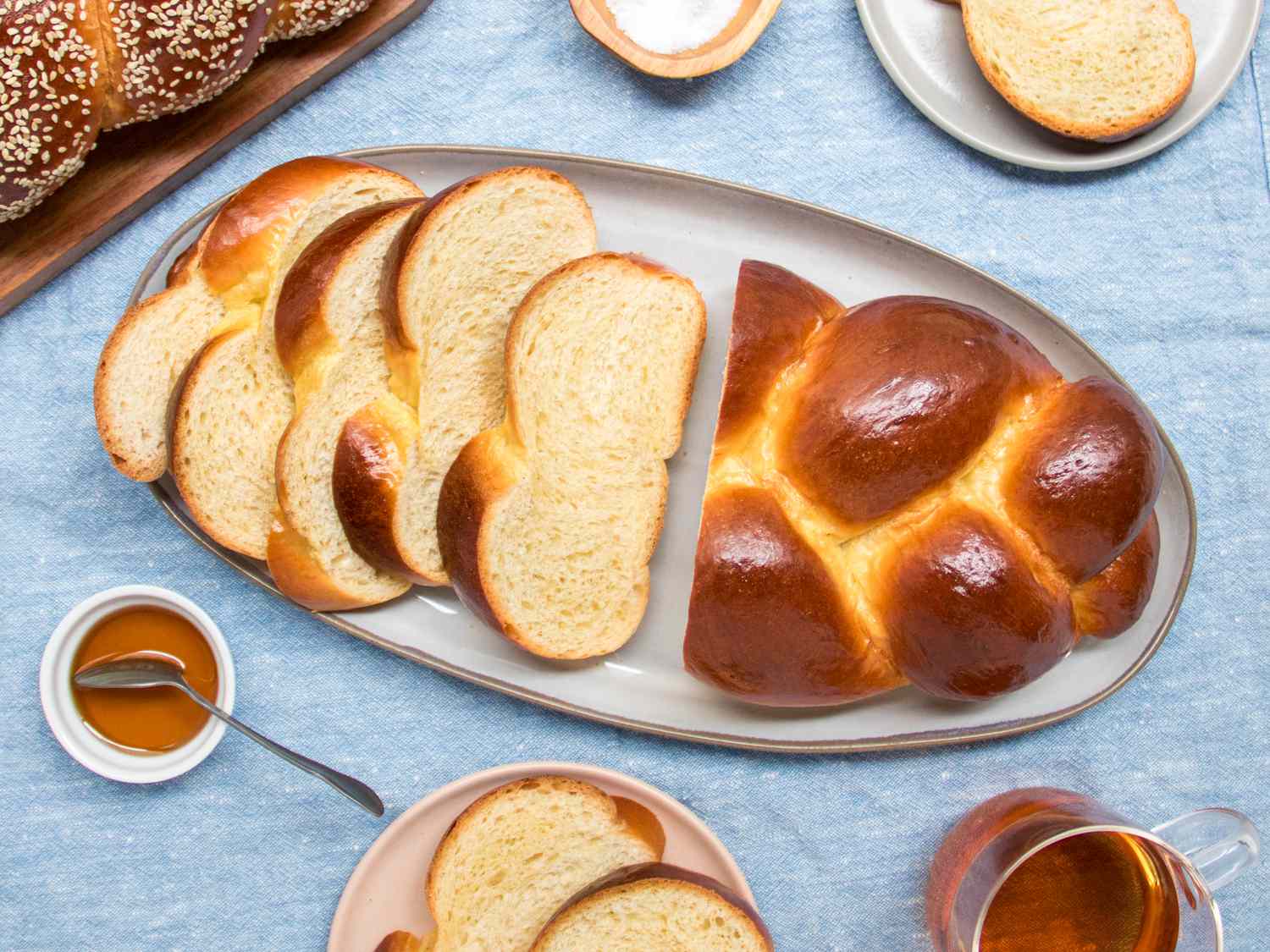 Entire loaf of plain woven challah on a plate, surrounded by smaller plates, salt, honey and a cup of tea