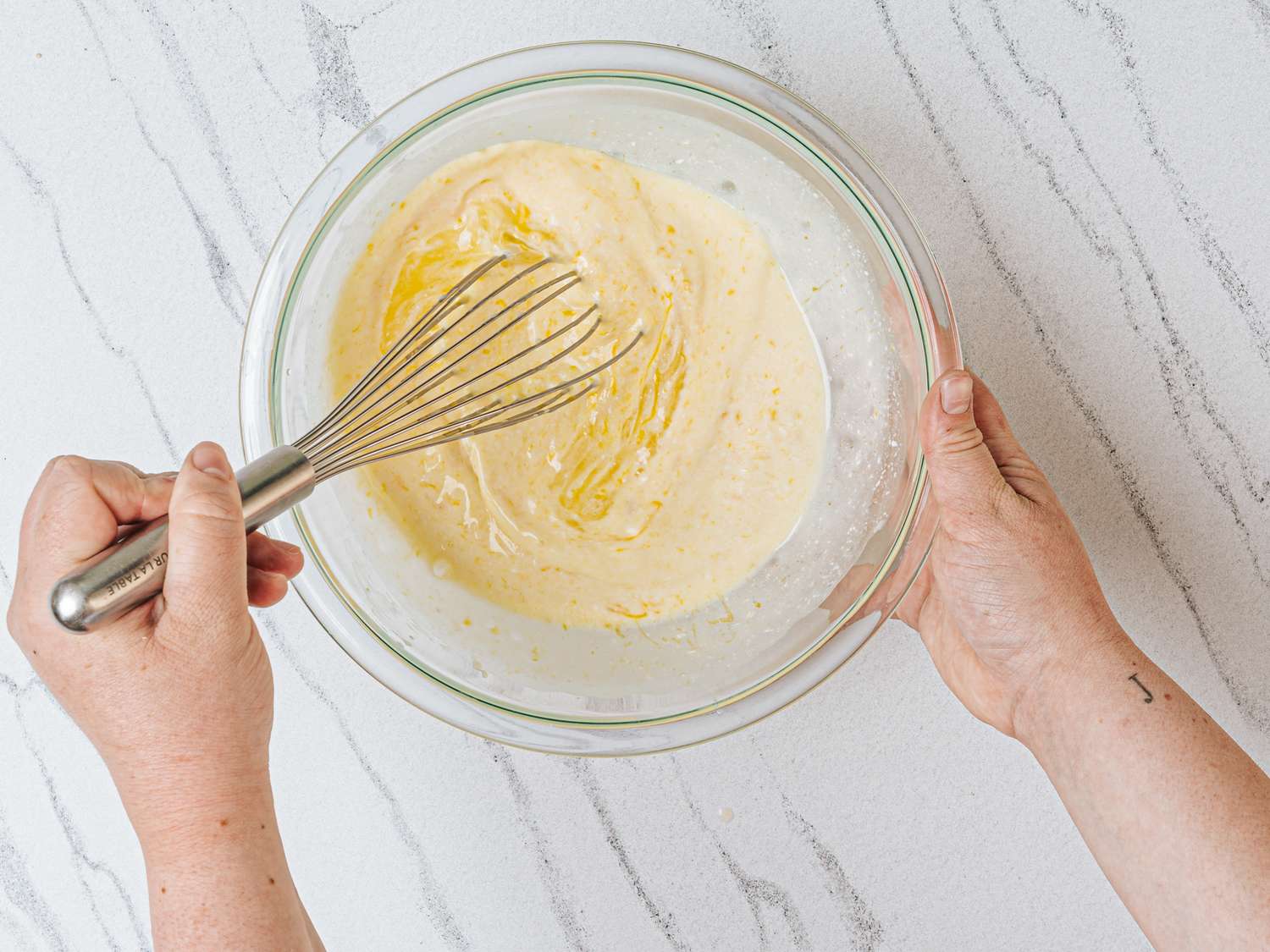 Two hands whisking a creamy yellow mixture in a glass bowl placed on a textured countertop