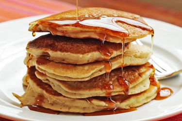 A stack of five pancakes on a plate with syrup being drizzled over them. 