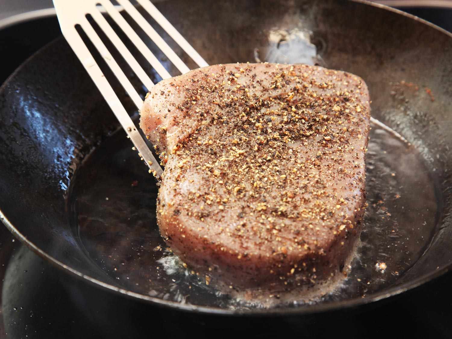 A tuna steak coated in coarsely ground black pepper being lowered into a preheated, oiled carbon steel skillet with a fish spatula.