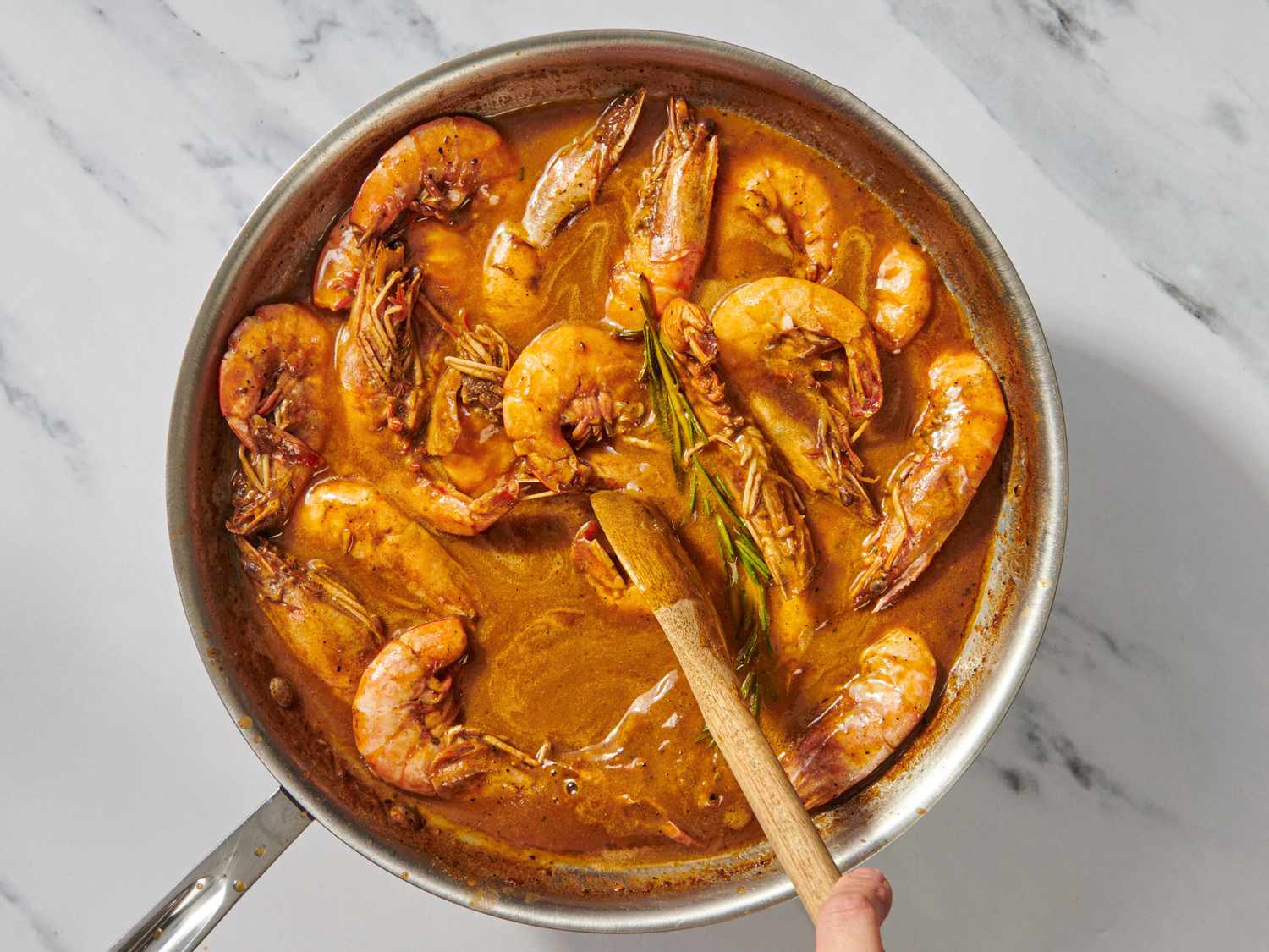 A skillet of New Orleansstyle BBQ shrimp with a reddish sauce and a sprig of rosemary being stirred with a wooden spoon