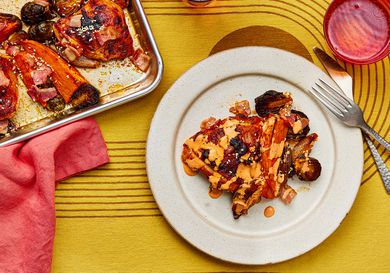 Serving of sheet-pan chicken and vegetables on a plate with a fork and knife and a red cup. To the side of the dish is the tray of chicken and vegetables with a red napkin