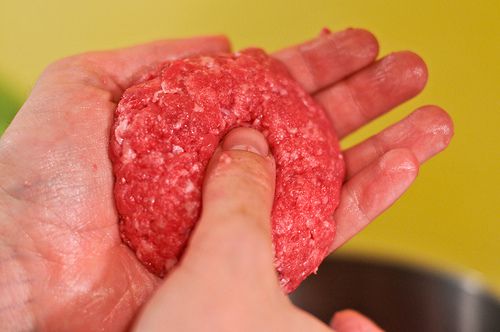 A hand shaping raw ground beef into a patty and using their thumb to make an indentation in the center.