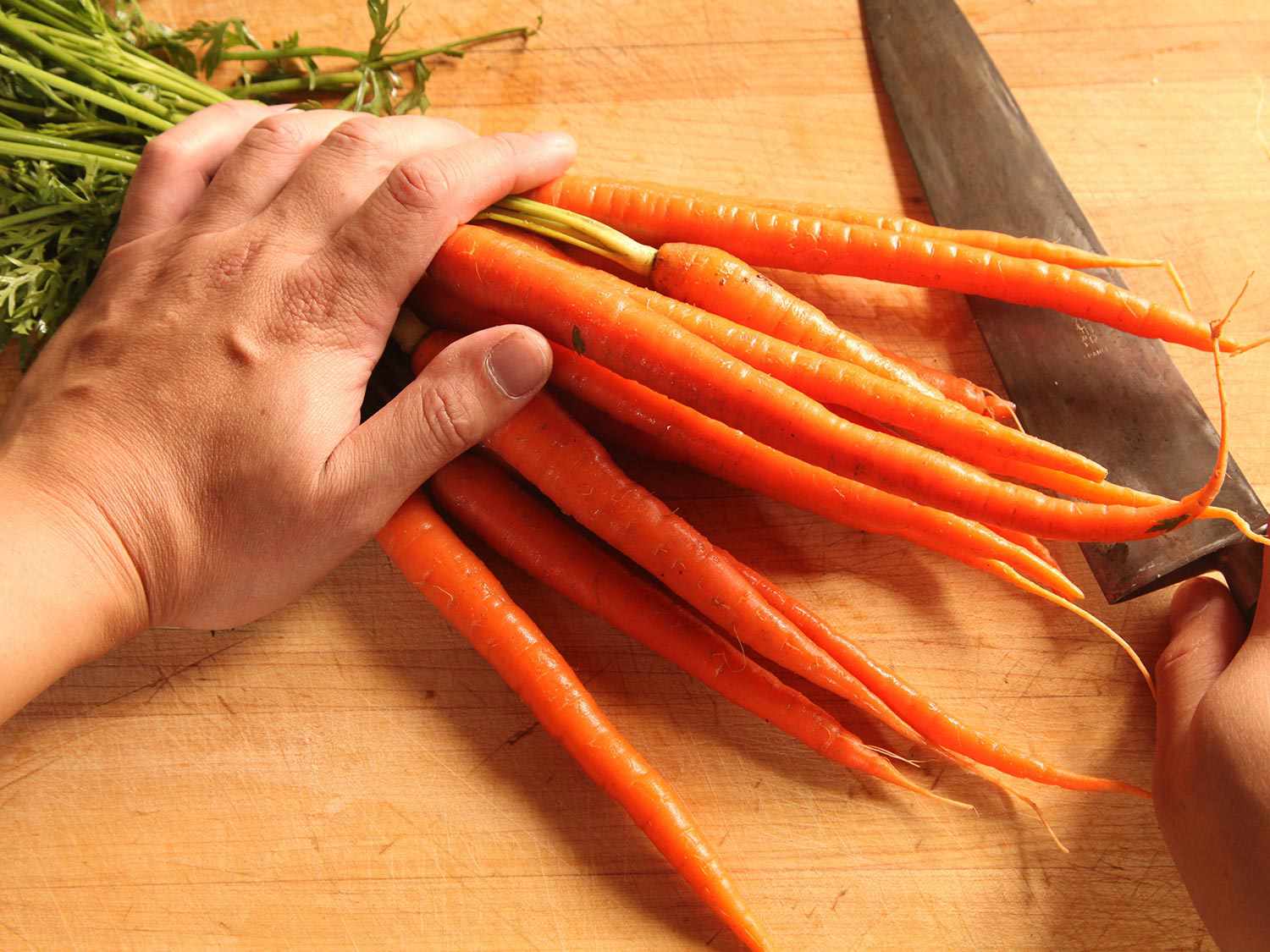 Hand pressing carrot bunch to cutting board, knife in other hand.