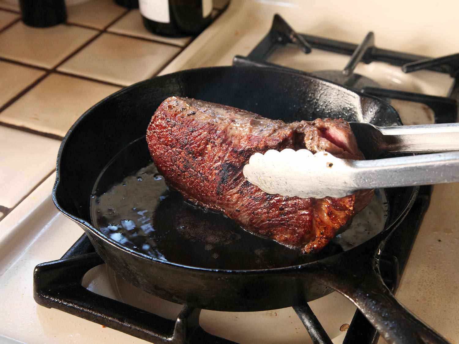 A cooked steak being turned on its edges by tongs to cook the sides on a cast iron skillet.