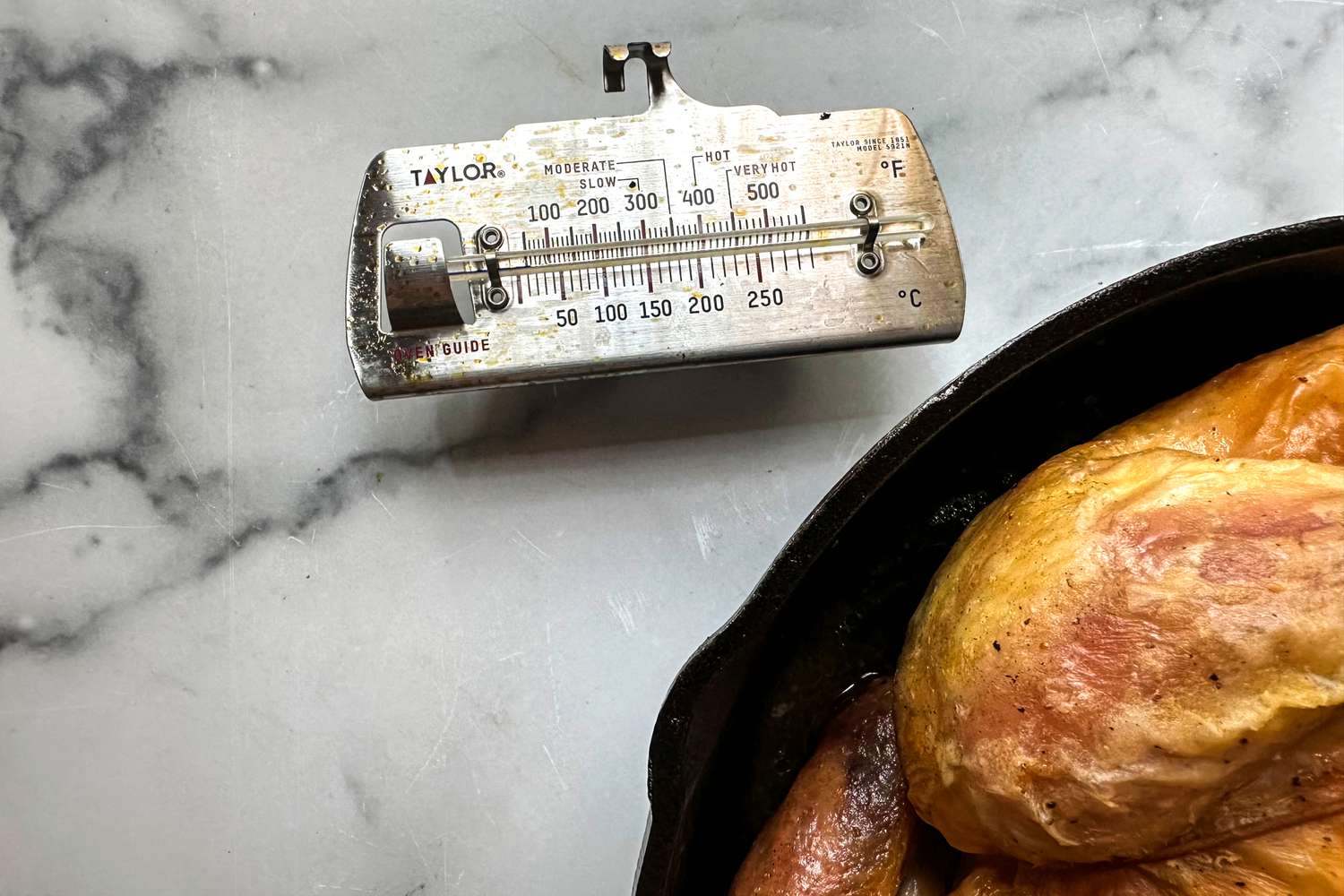 A metal analog oven thermometer on a marble counter next to a cast iron pan.