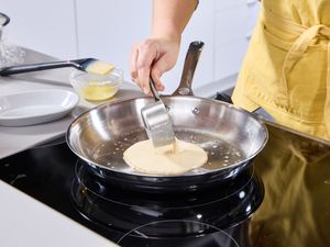 A person pouring crepe batter into a stainless steel skillet