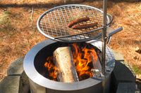Hot dogs cooking on a grill over a flaming outdoor firepit