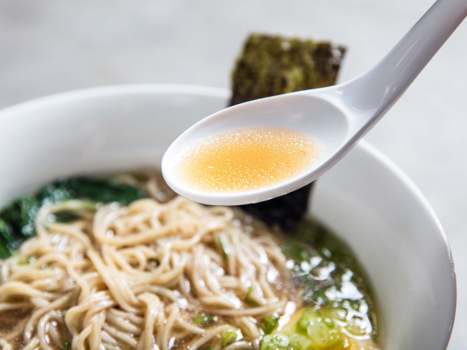 A spoonful of shoyu ramen broth being lifted from a bowl of broth and noodles.