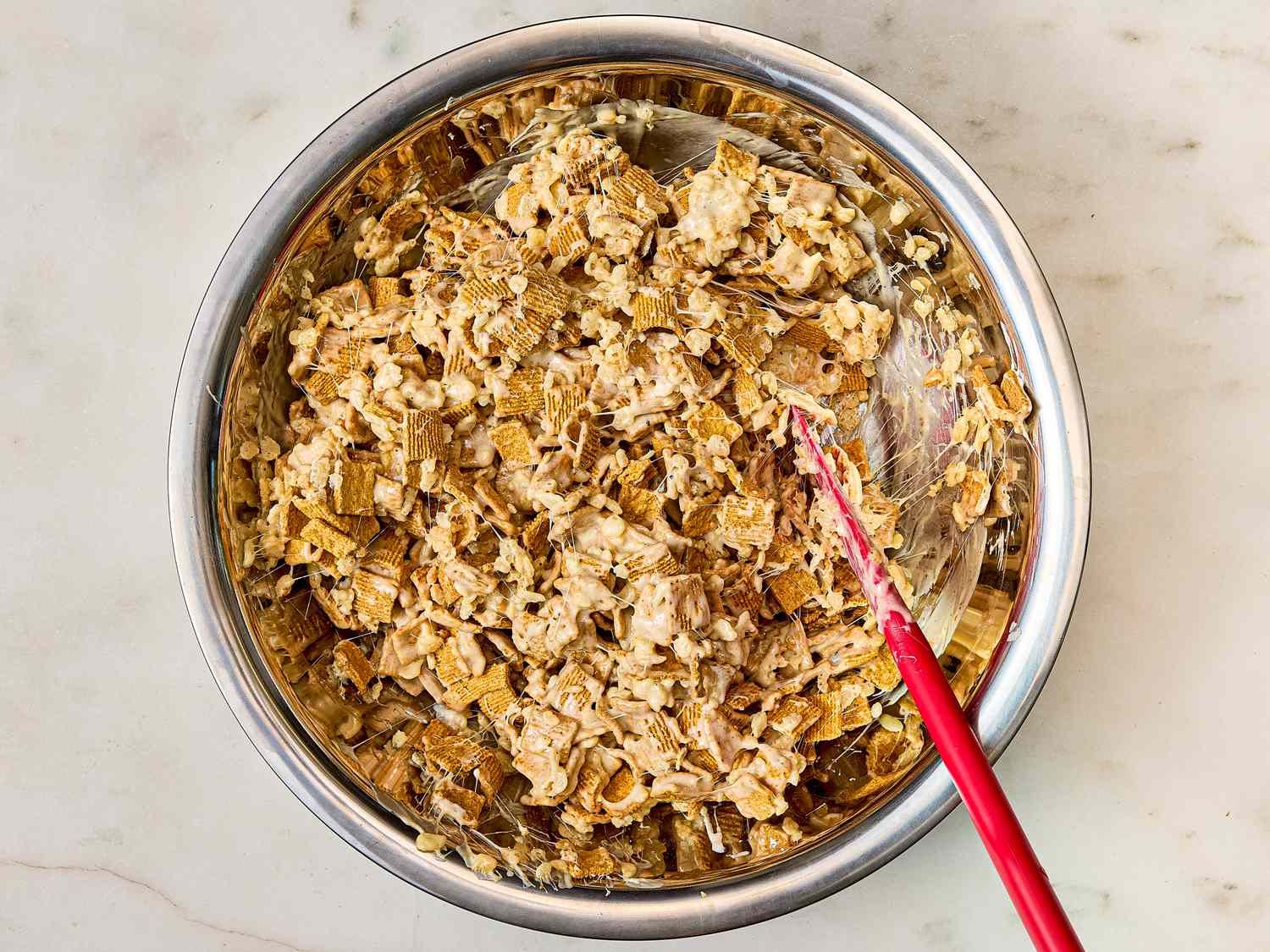 A mixing bowl with a marshmallow and cereal mixture being stirred with a red spatula