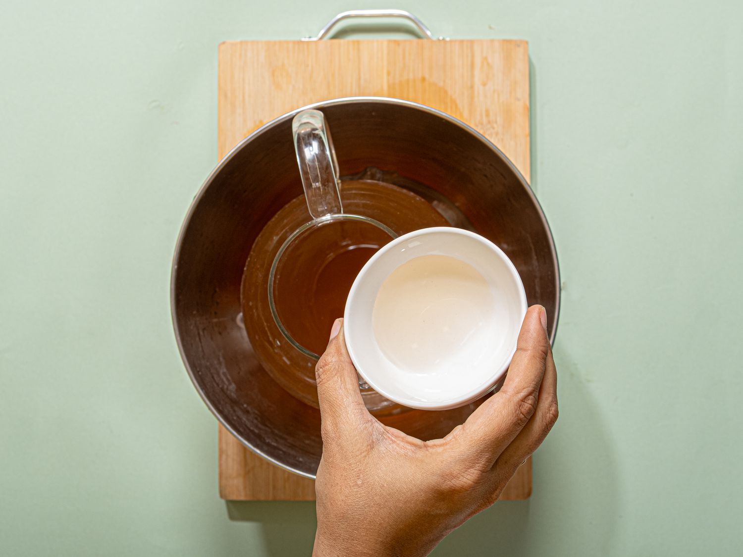 A hand holding a bowl above a mixing bowl on a wooden board
