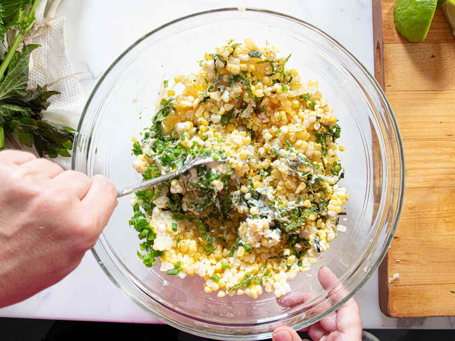 A bowl of corn salad being mixed with a fork hands visible holding the bowl