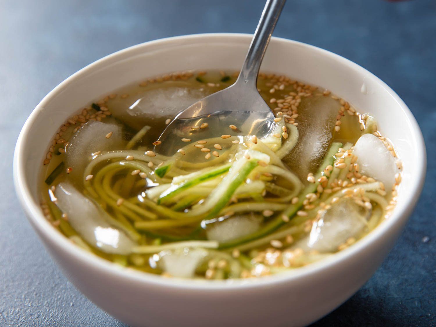 A spoon sips into a bowl of oi naengguk, an icy chilled korean soup, will ice cubes floating in the bowl with the cucumbers and broth
