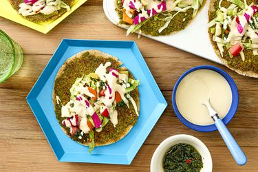 A smash falafel pita dressed with toppings next to more smash falafel pitas, a bowl of tahini sauce, and a bowl of zhug. 