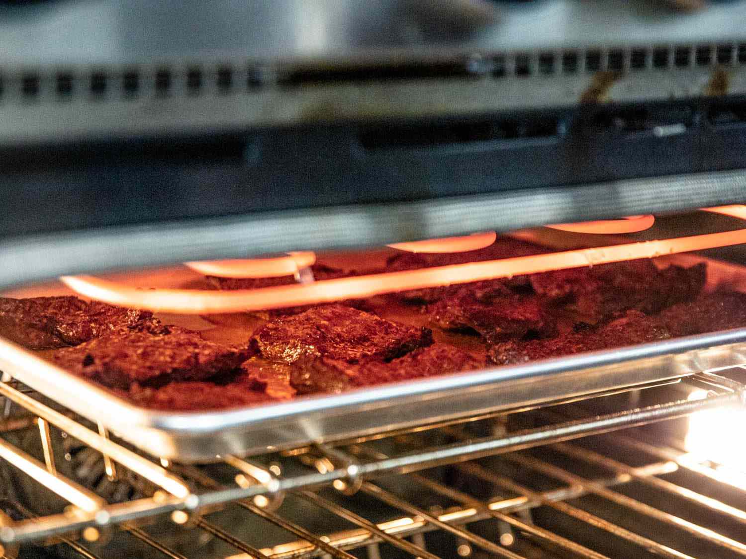 Skirt steaks on a rimmed sheet pan under an oven's hot broiler element; the oven rack is in its highest position