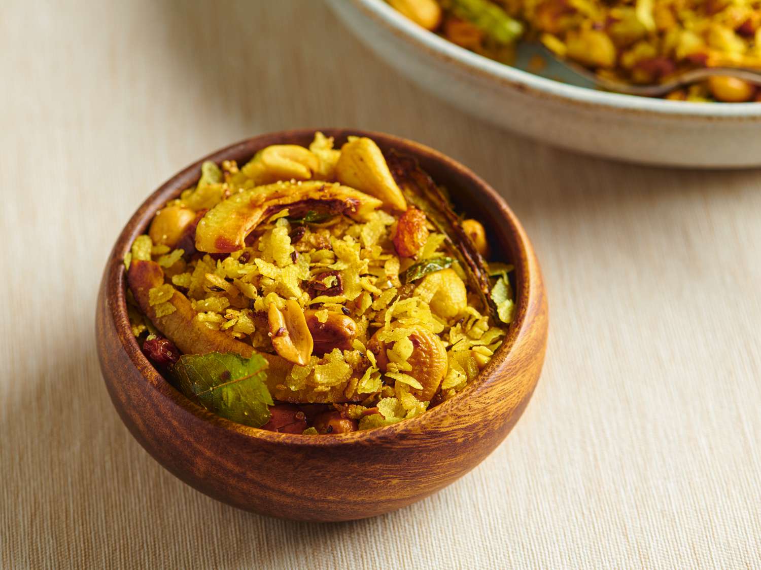 A small round wooden bowl holding poha chivda. There is a larger cream-colored ceramic bowl in the top right corner of the image.