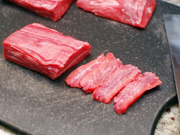 A slab of flank steak, half cut into thin rectangular slices on a black surface. 