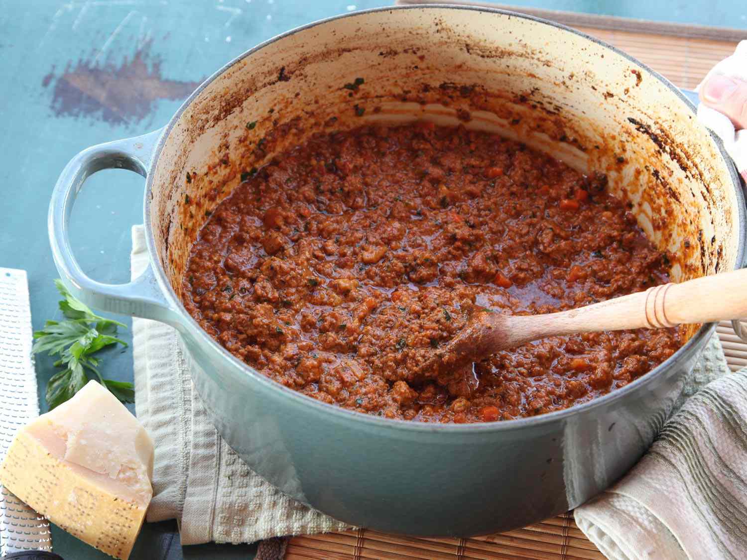 Meat sauce in a blue Dutch oven being stirred with a wooden spoon with a cut of Parmesan cheese resting off to the side.