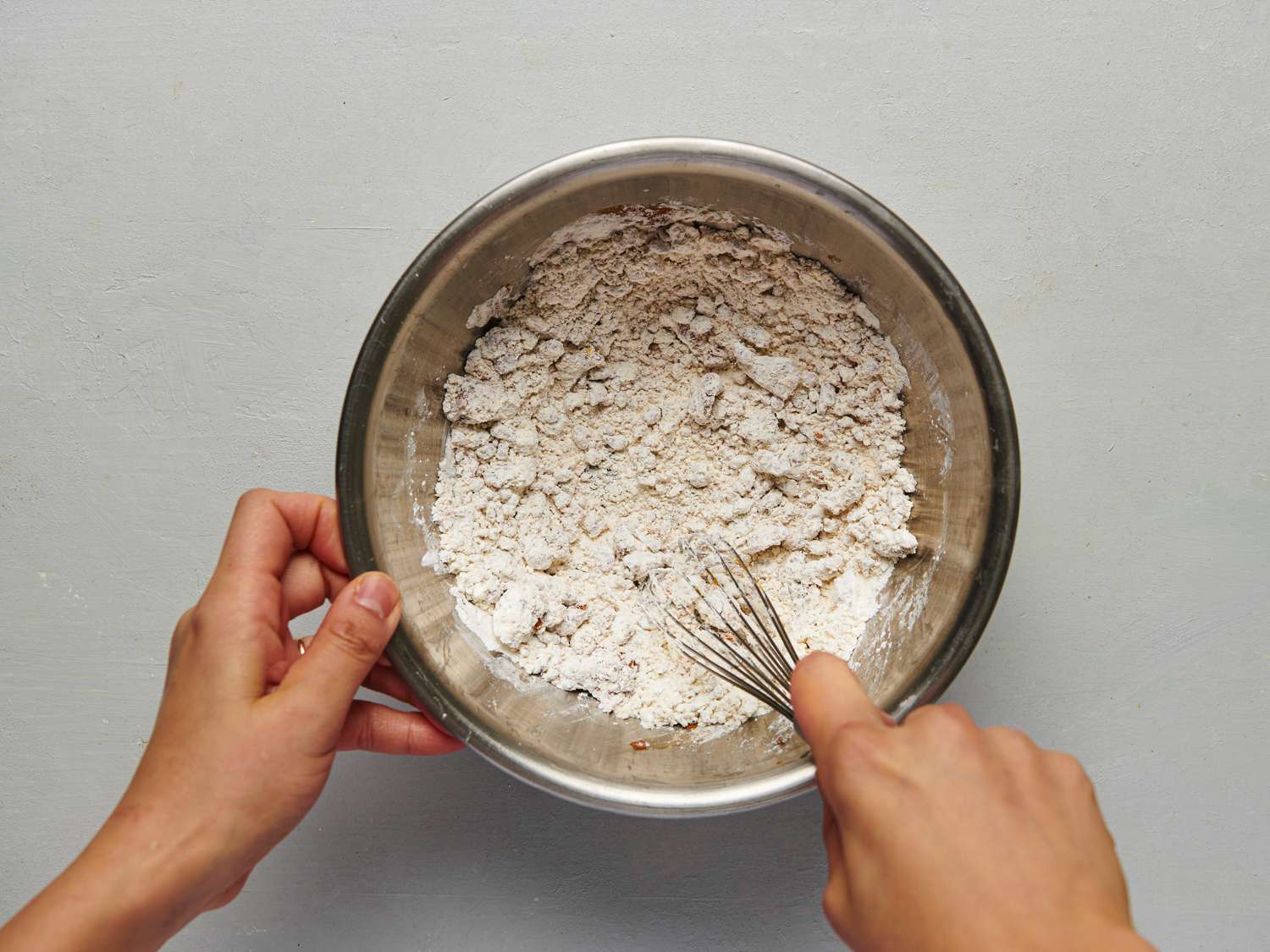 Reserved marinade whisked into the dry mixture, showing off coarse, mealy clumps. There is a pair of hands holding a whisk which is inside of the bowl.