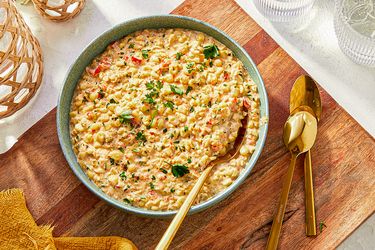 A bowl of creamy corn garnished with herbs on a wooden surface with golden utensils alongside