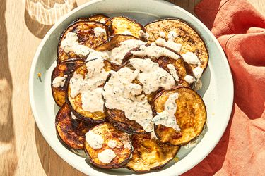 Grilled eggplant slices topped with a creamy white sauce served in a bowl on a wooden table beside a red cloth napkin