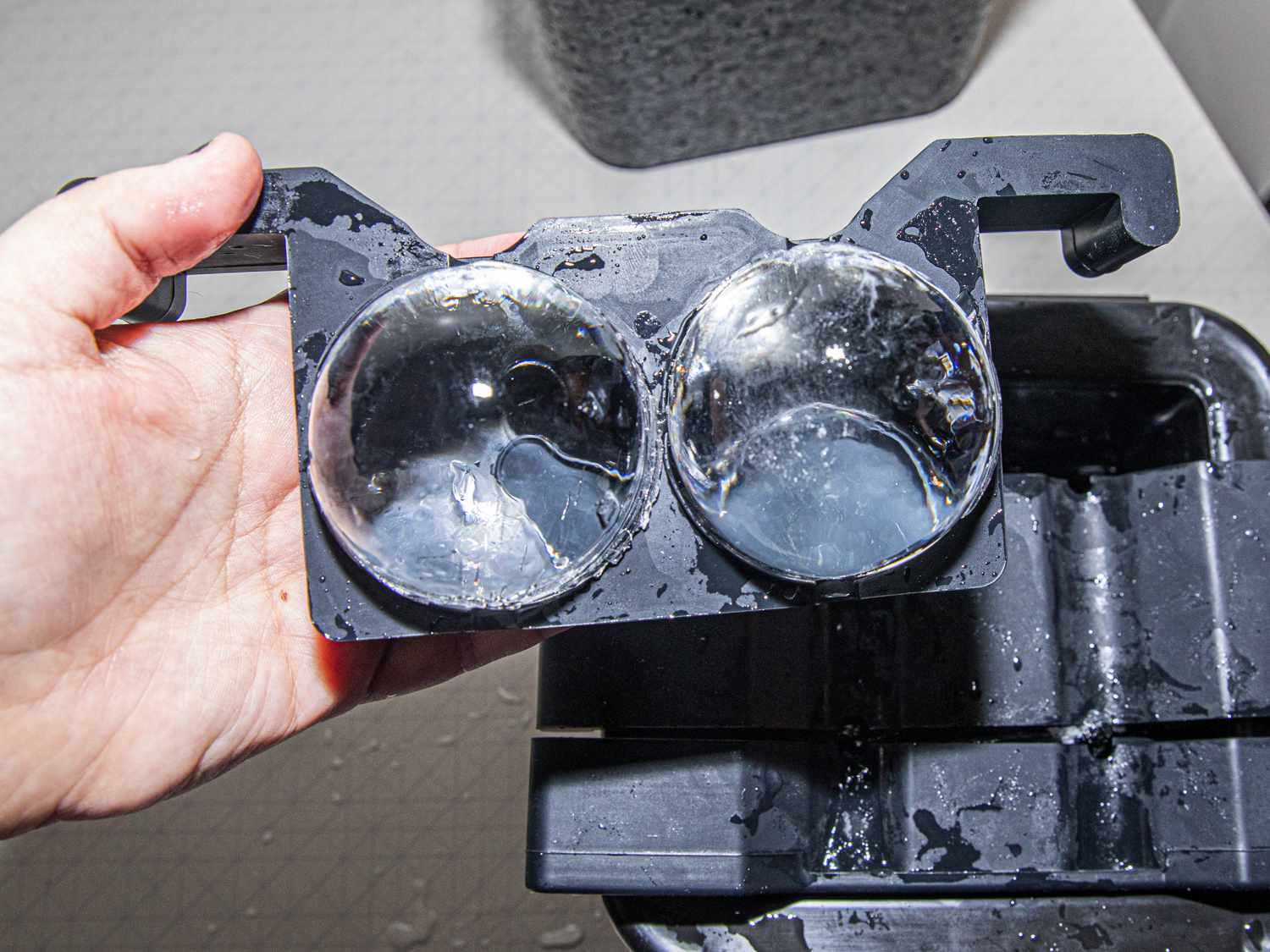 Two clear ice balls in their black plastic tray with a hand holding the tray up