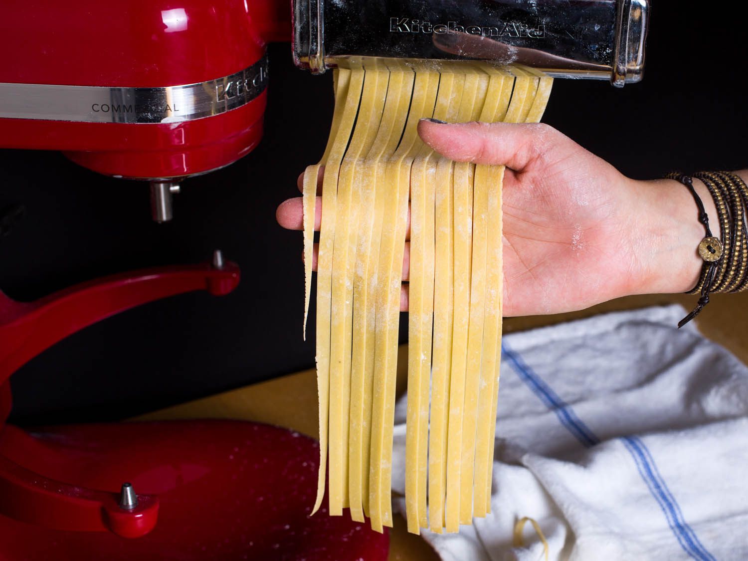 A hand receiving cut fresh fettuccine as it comes out the end of a pasta-cutting attachment on a stand mixer.