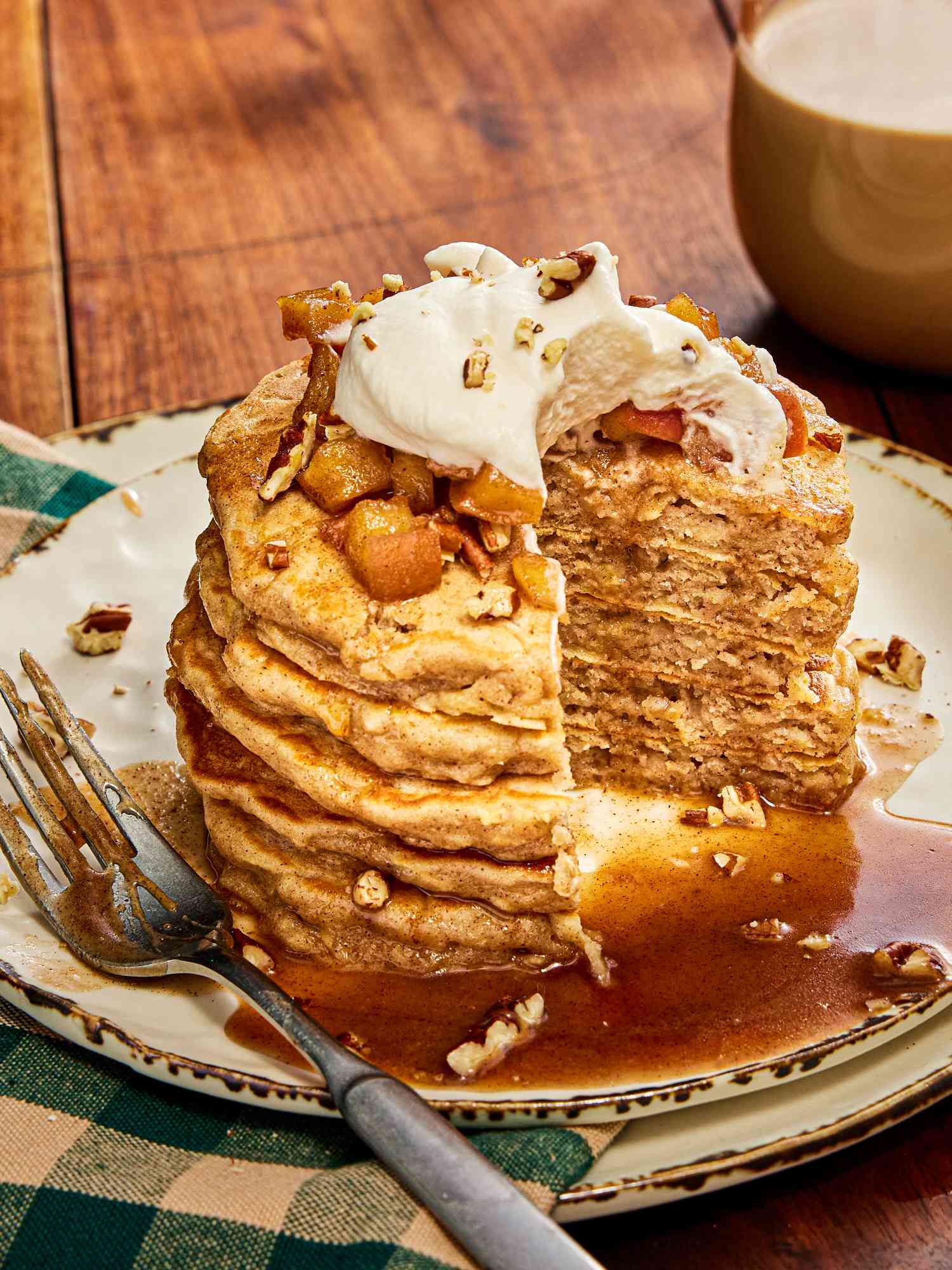 Stack of apple pie pancakes with a portion taken out, on white rustic plates, a green checked napkin, and warm drink
