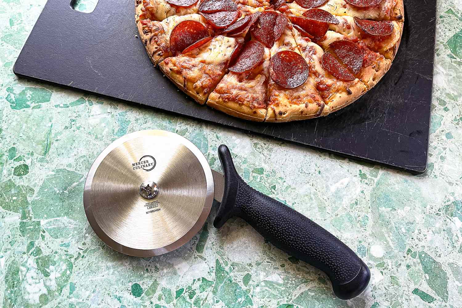 A pepperoni pizza partially sliced on a cutting board lying next to a Mercer pizza cutter with a textured handle on a green marble countertop