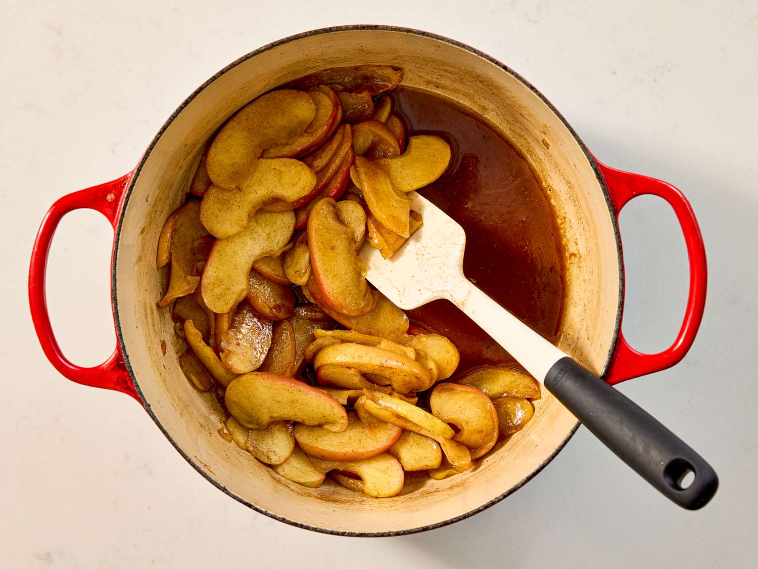 Cooked apple slices in a red pot with a spatula preparation for a caramel tart
