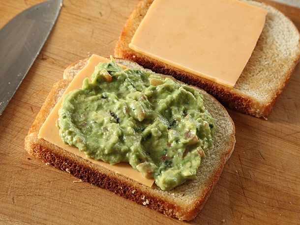 Two slices of bread, with one topped with a slice of cheddar and the other with cheddar and a thick spread of guacamole.
