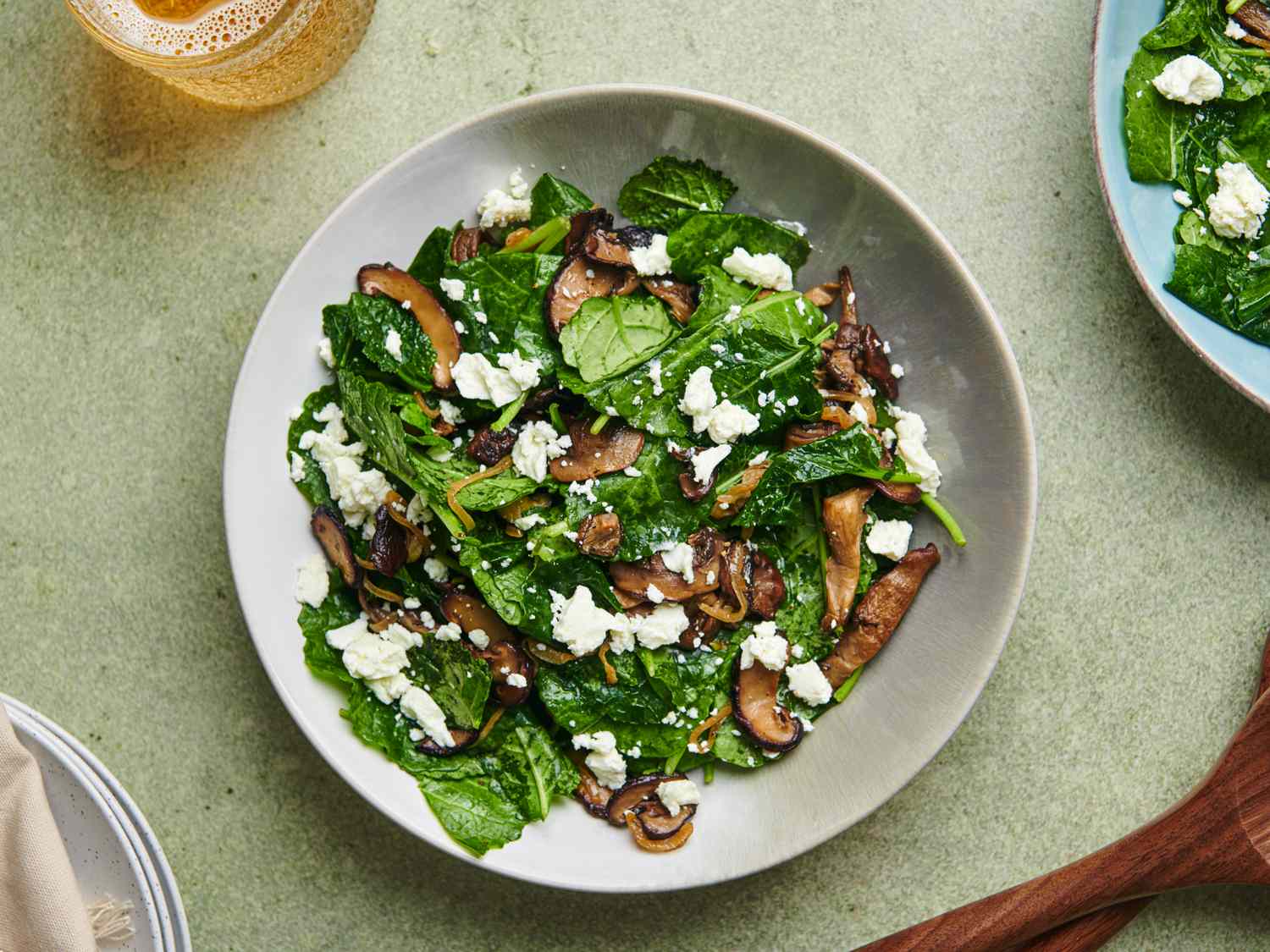 Warm kale and caramelized mushroom salad in a white ceramic bowl on a green surface. There are a pair of wooden spoons at the bottom right corner, a glass of golden liquid in the top left corner, a stack of plates covered with a napkin in the bottom left corner, and another plate of salad in the top right corner.