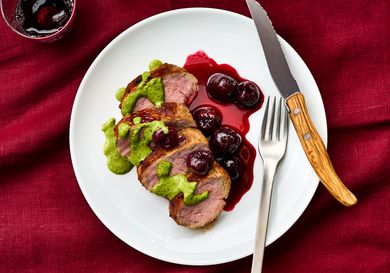 Sliced roasted pork tenderloin with green sauce and roasted cherries on a plate, placed on a red tablecloth with a fork and knife