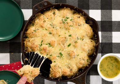 pullinh cheese garlic knot from cast iron pan and small dish of garlic butter on a black and white checkered fabric. 2 green plates and red napkin to the sides. 