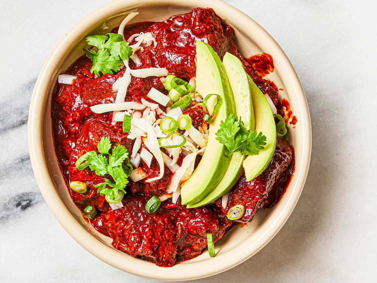 Overhead view of finished chili con carne in a bowl topped with scallions, cheese, fresh cilantro, and avocado slices.