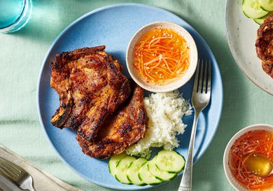 Pork chops over steamed rice white on a plate with sliced cucumbers and a ramekin of sauce