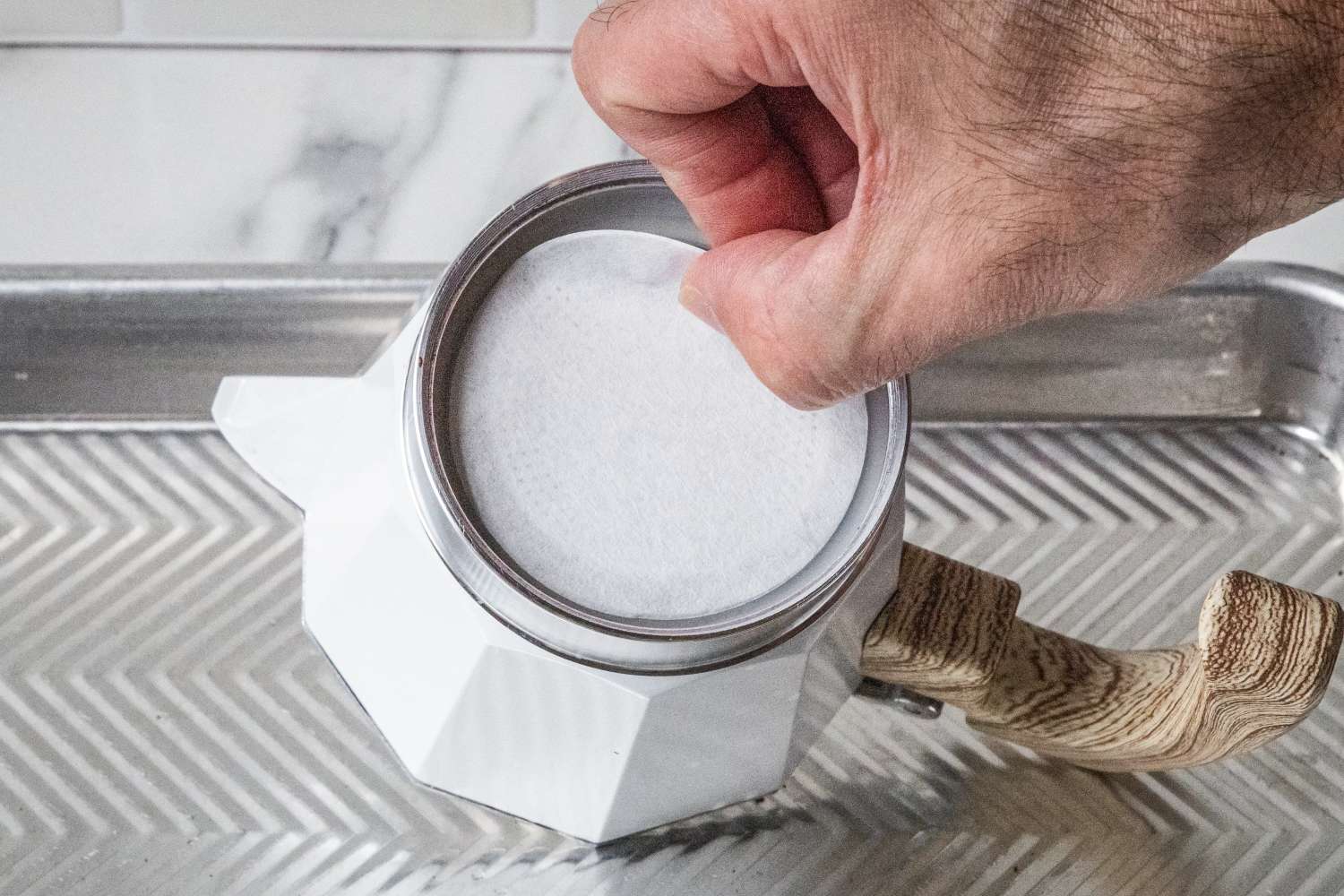 A hand placing a circular filter into the top of a coffee pot with a wooden handle positioned on a textured metal surface