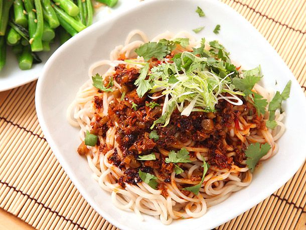 A dish of vegan dan dan noodles topped with a savory mix of sauce, garnishes, and herbs, served on a bamboo mat near a side of green vegetables