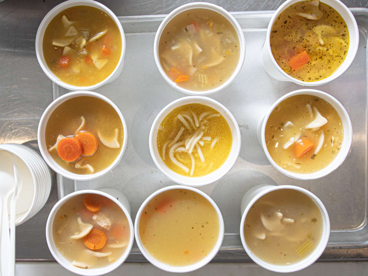 Nine cups of chicken noodle soup arranged on a tray showcasing ingredients like noodles carrots and broth