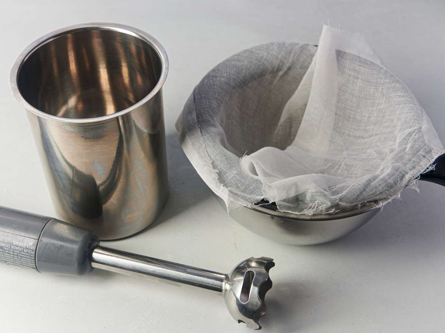 Tall and narrow vessel set up with immersion blender, and a fine mesh strainer covered with cheesecloth and set over a heatproof bowl