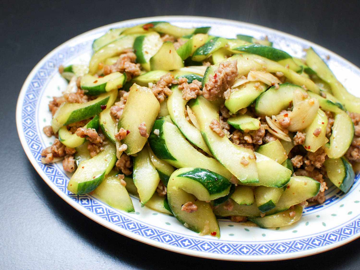 A serving dish piled high with stir-fried cucumbers and ground pork, flecked with slices of garlic and chile flakes.