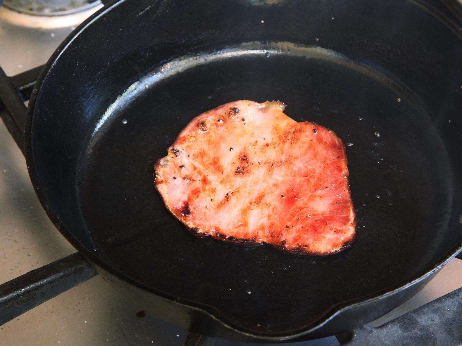 A slice of Canadian bacon in a pan, ready to go on a homemade Egg McMuffin