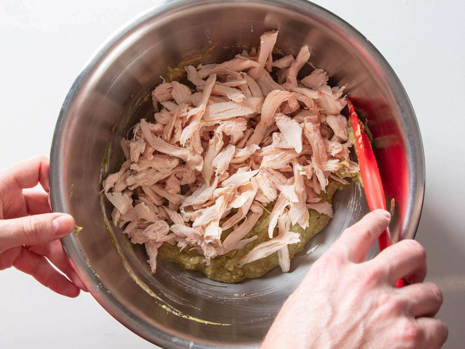 Tossing shredded chicken with pesto dressing in a mixing bowl.